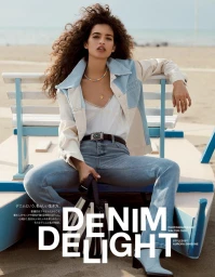 Woman with long curly brown hair wearing a patchwork denim jacket, white camisole, and light blue jeans, seated on a white wooden bench on a sandy beach.