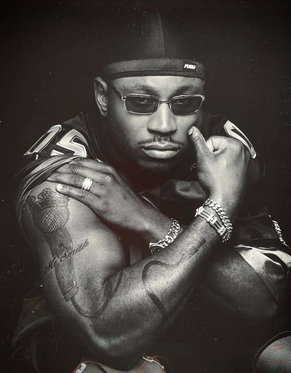 A black and white studio portrait of an adult male wearing sunglasses, chains, and a durag looking at the camera with serious expression.