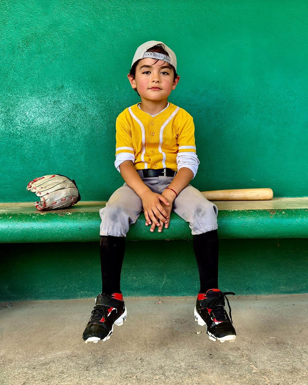 Young boy in baseball uniform sits on bench with baseball bat and glove in dugout, wearing cap and black baseball socks.