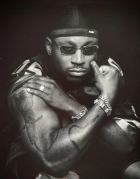 Portrait of LL Cool J wearing a dark durag, sunglasses, and jersey, with arms crossed displaying tattoos and silver bracelets.