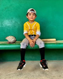 Young child in a yellow baseball jersey and cap sitting on a green bench with a glove and bat.
