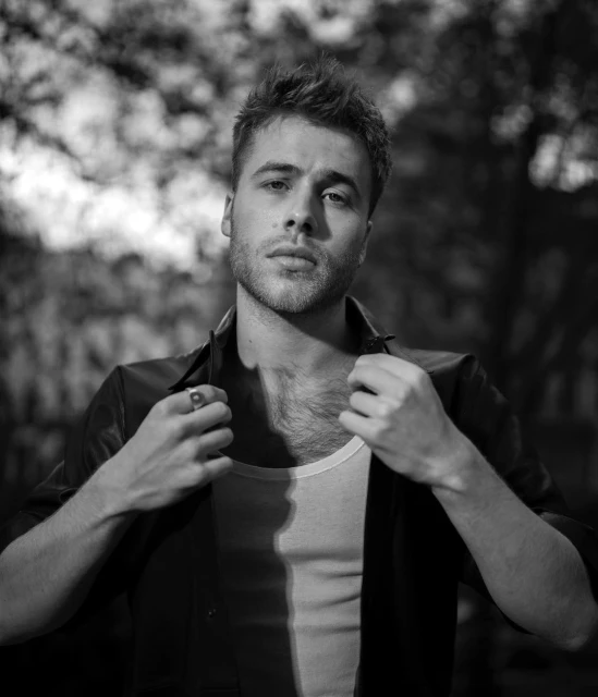Black and white portrait of a young adult man in a leather jacket, gazing at the camera with a neutral expression against a blurry outdoor backdrop.