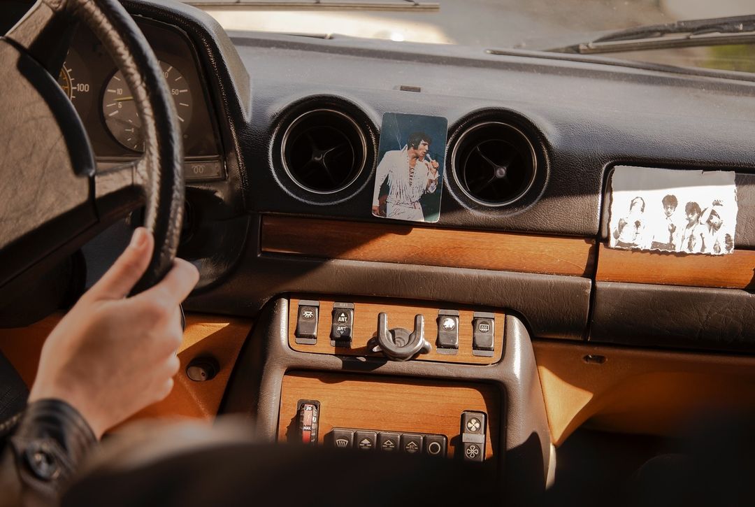 Car interior with steering wheel, dashboard, Elvis Presley photo, and group picture, showcasing a vintage vehicle's design and driver's touch.