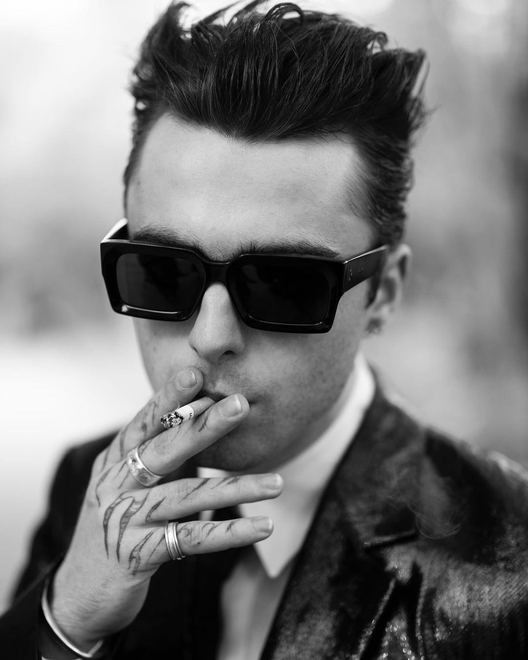 Black and white portrait of an adult male wearing dark glasses and a suit, smoking a cigarette, with tattoos on his fingers.