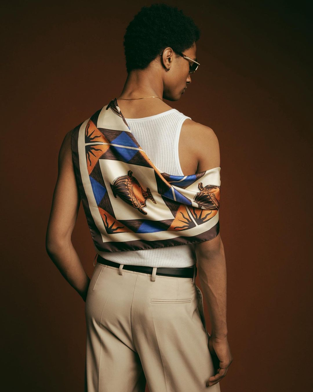 Back view of a man with a patterned scarf and glasses posing against a brown background in a studio, fashion-forward and stylish.