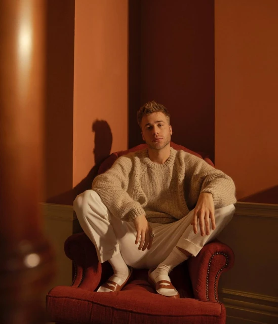 Blond male model sits cross-legged in a red armchair, wearing a cream sweater and white pants, posing in a warm-toned interior space.
