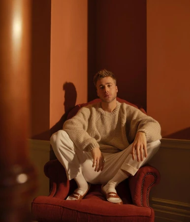 Blond male model sits cross-legged in a red armchair, wearing a cream sweater and white pants, posing in a warm-toned interior space.
