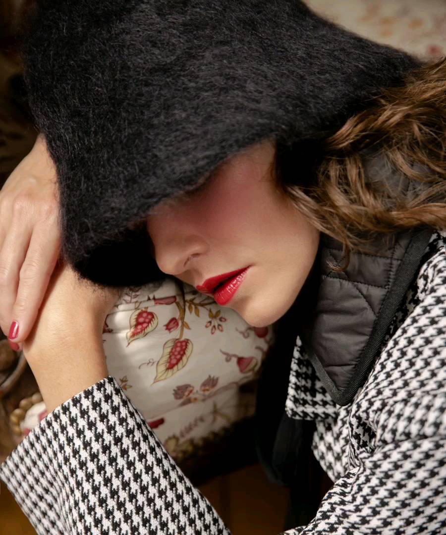 Close-up portrait of a woman with dark, fluffy hat, red lipstick, resting her face on a floral cushion and arm with black and white patterned jacket.
