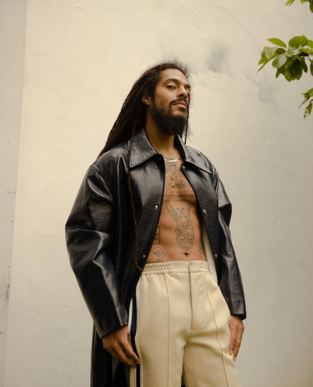 Man with dreadlocks poses in a black coat and cream pants against a white wall with green foliage in the upper corner.