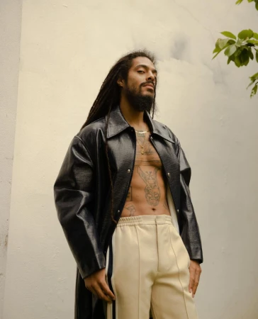 Man with dreadlocks poses in a black coat and cream pants against a white wall with green foliage in the upper corner.