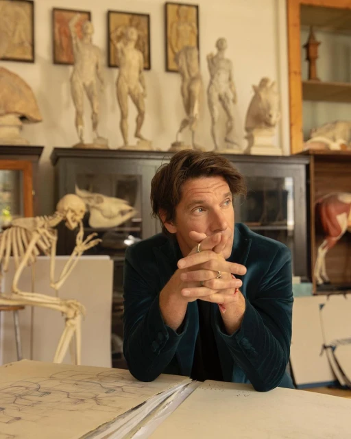 Adult male in a green velvet blazer seated indoors surrounded by drawing, anatomical models, and sculptures with natural window lighting.