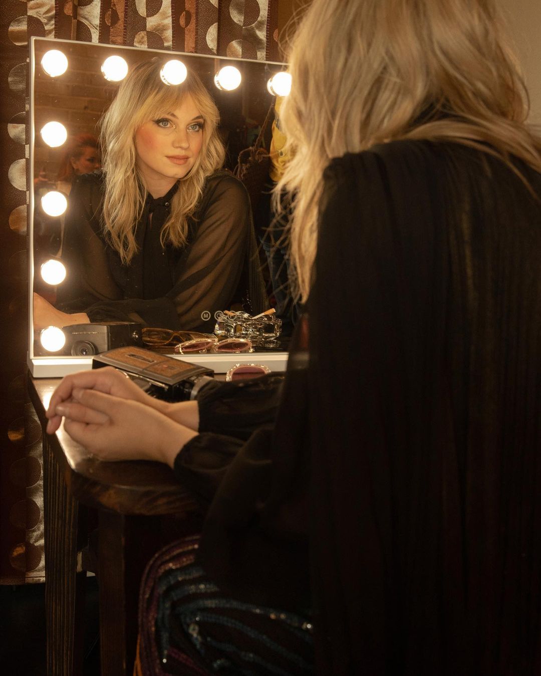Blonde woman sits at a vanity mirror with illuminated lights, applying makeup. She wears a black top and has a neutral expression.