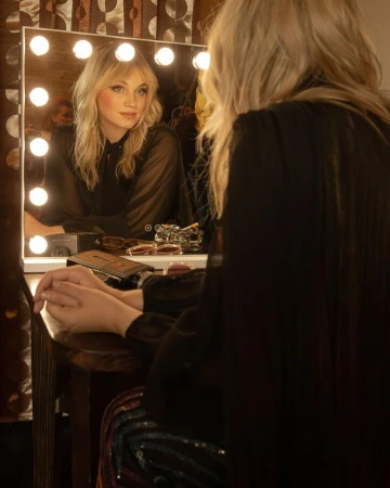 Blonde woman sits at a vanity mirror with illuminated lights, applying makeup. She wears a black top and has a neutral expression.