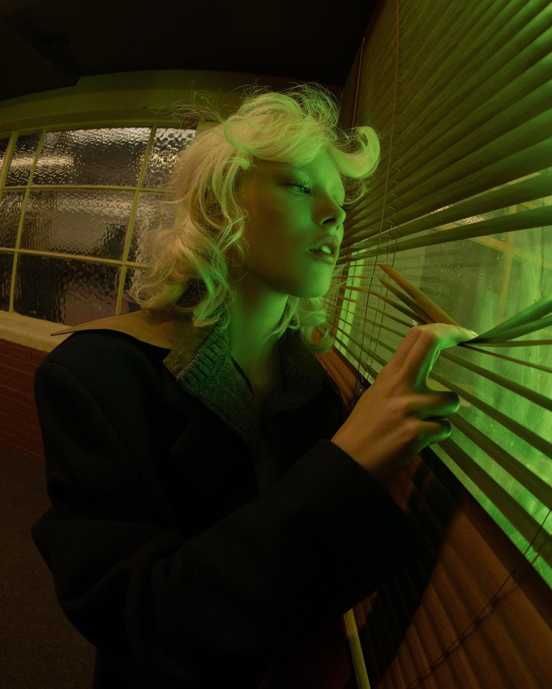 Indoor portrait of a young blonde woman in a coat looking through window blinds, illuminated by green light, creating a mysterious atmosphere.