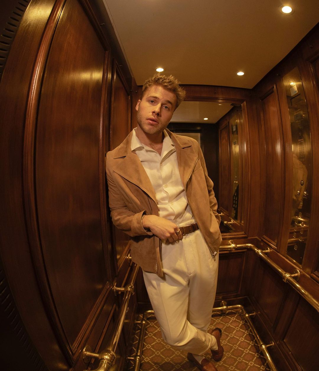 Portrait of a blond male in a tan jacket and white pants, posing inside an elevator with wood paneled walls and a patterned floor.
