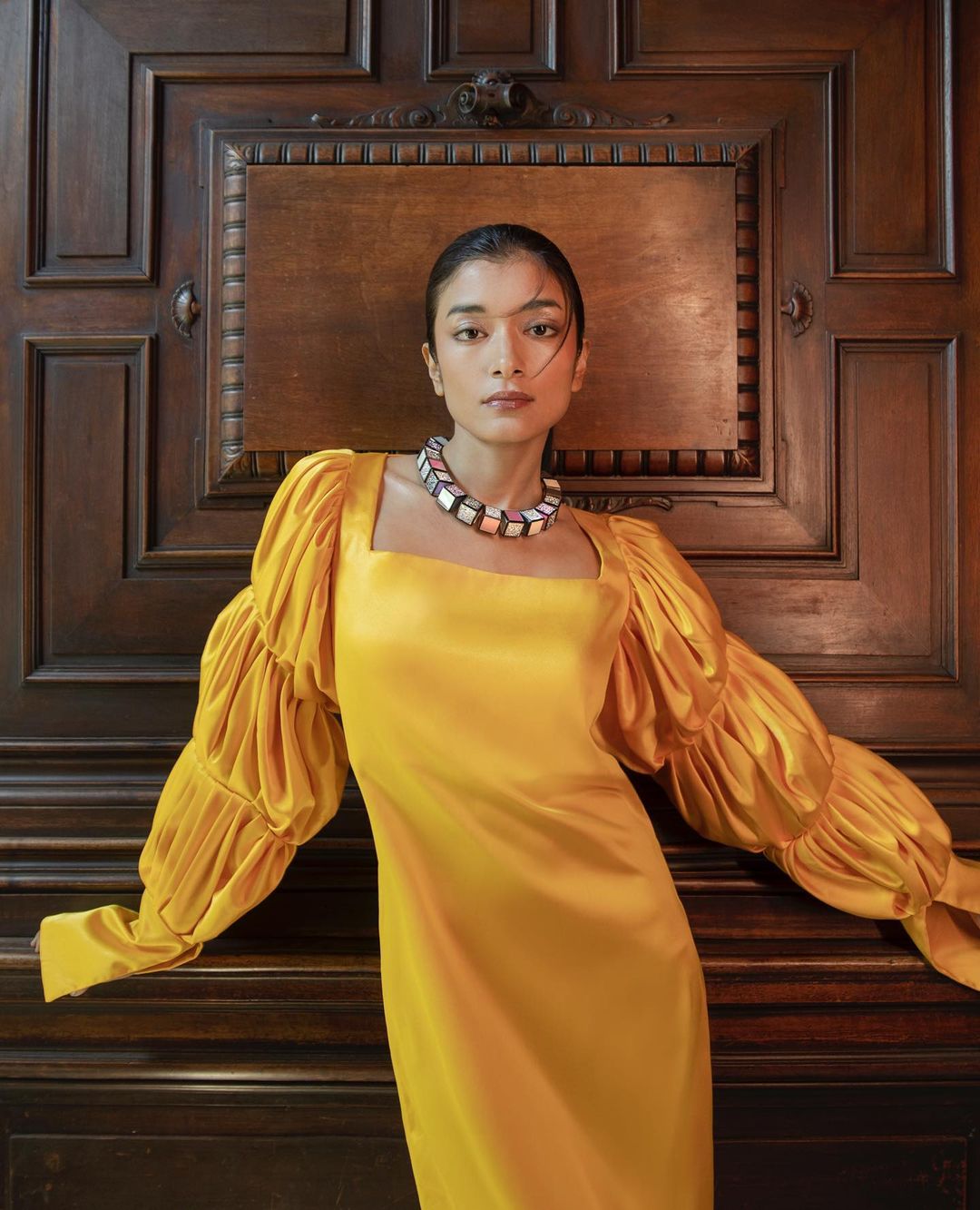Elegant woman in a yellow dress poses against a paneled wood wall. She wears a bold necklace and has a serene facial expression.