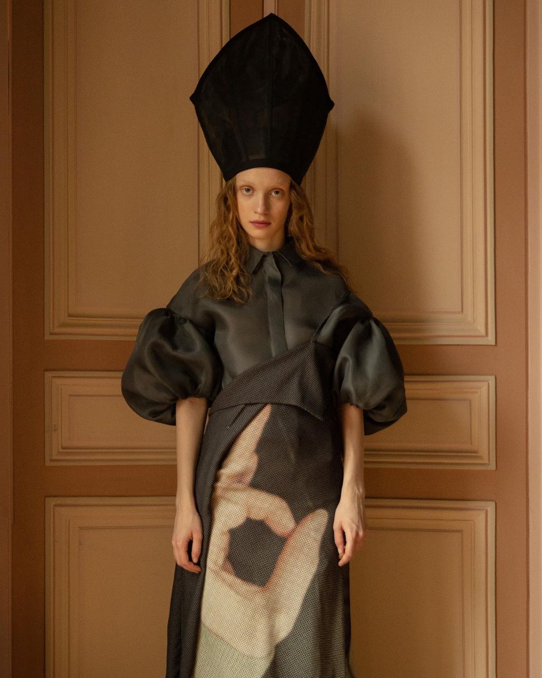 A model wearing an extravagant avant-garde black hat and fashion dress, posing indoors with a neutral color door in the background.