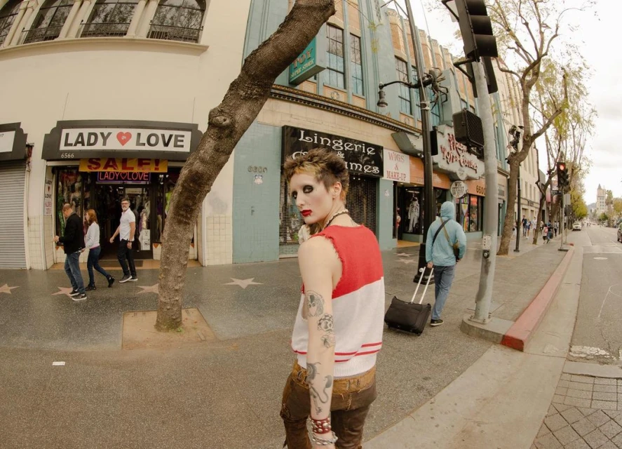A person stands on a sidewalk in an urban setting, wearing a red and white top and brown pants, with shops and pedestrians nearby.