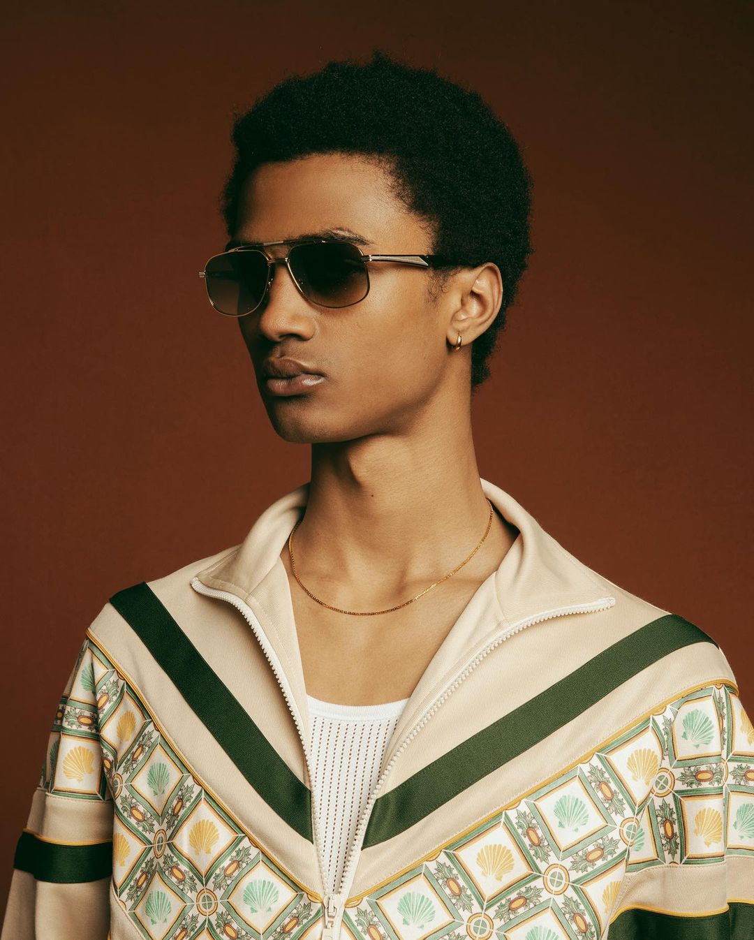 Headshot of a Black male model wearing aviator sunglasses and a patterned jacket against a brown background in a studio setting.