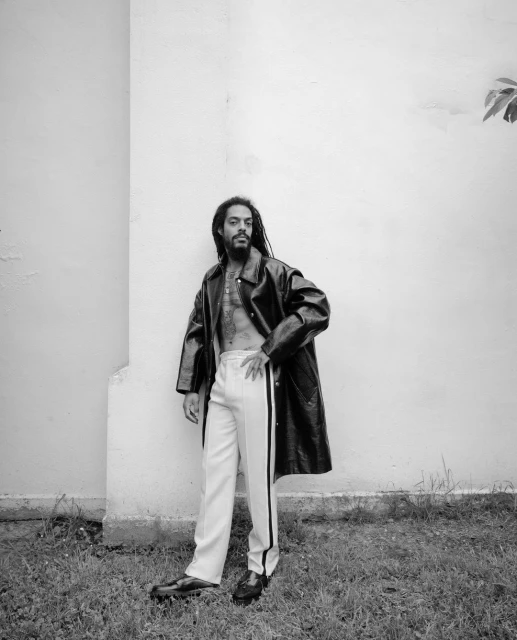 Black and white portrait of a man with dreadlocks wearing a leather coat and track pants, posing against a light wall outdoors.