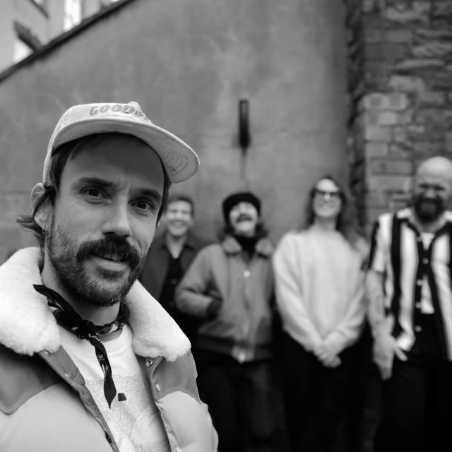 Black and white shot of five people outside in a city setting, featuring one adult male in the foreground wearing a cap.