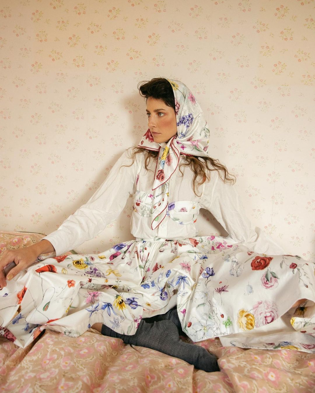 Adult woman in floral headscarf and dress sitting on a bed in front of floral wallpaper, looking away in a fashion pose.