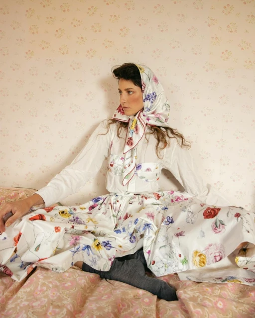 Adult woman in floral headscarf and dress sitting on a bed in front of floral wallpaper, looking away in a fashion pose.