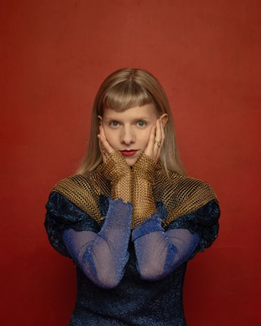 Close-up studio shot of a woman with fair skin, blonde hair with bangs, wearing a blue dress and gold mesh sleeves, looking at camera.