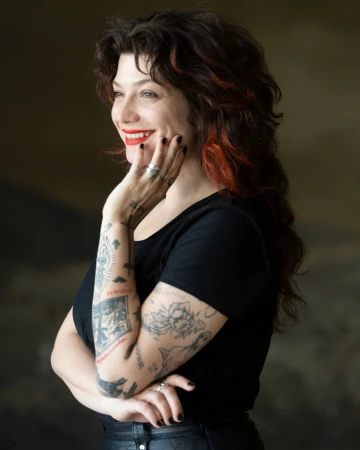 Portrait of a tattooed woman with dark curly hair wearing a black top and smiling with hand to cheek, by photographer Wanda Martin.