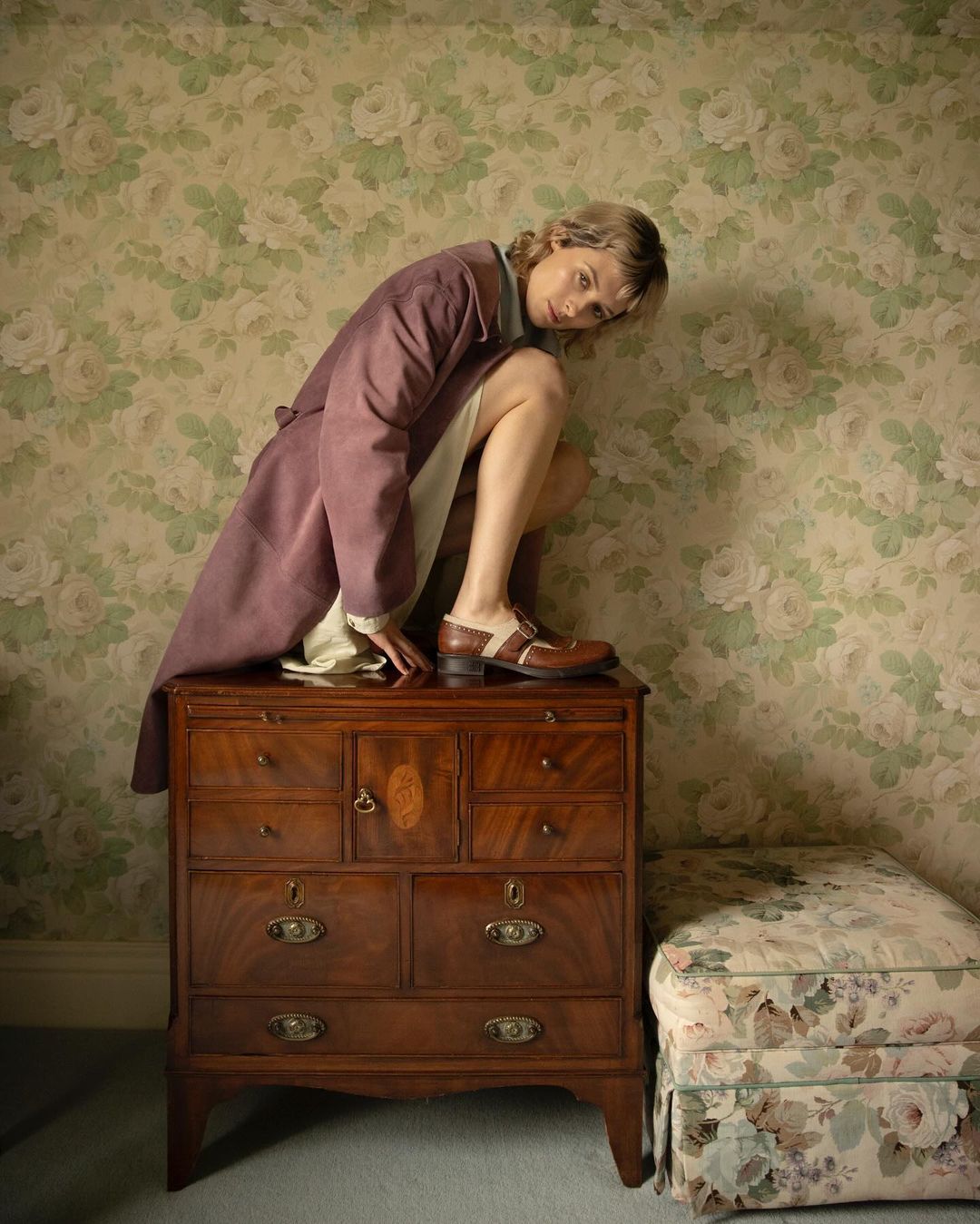 Young woman in a coat poses atop a wooden dresser, showcasing a quirky, vintage fashion sensibility against a floral wallpaper backdrop.