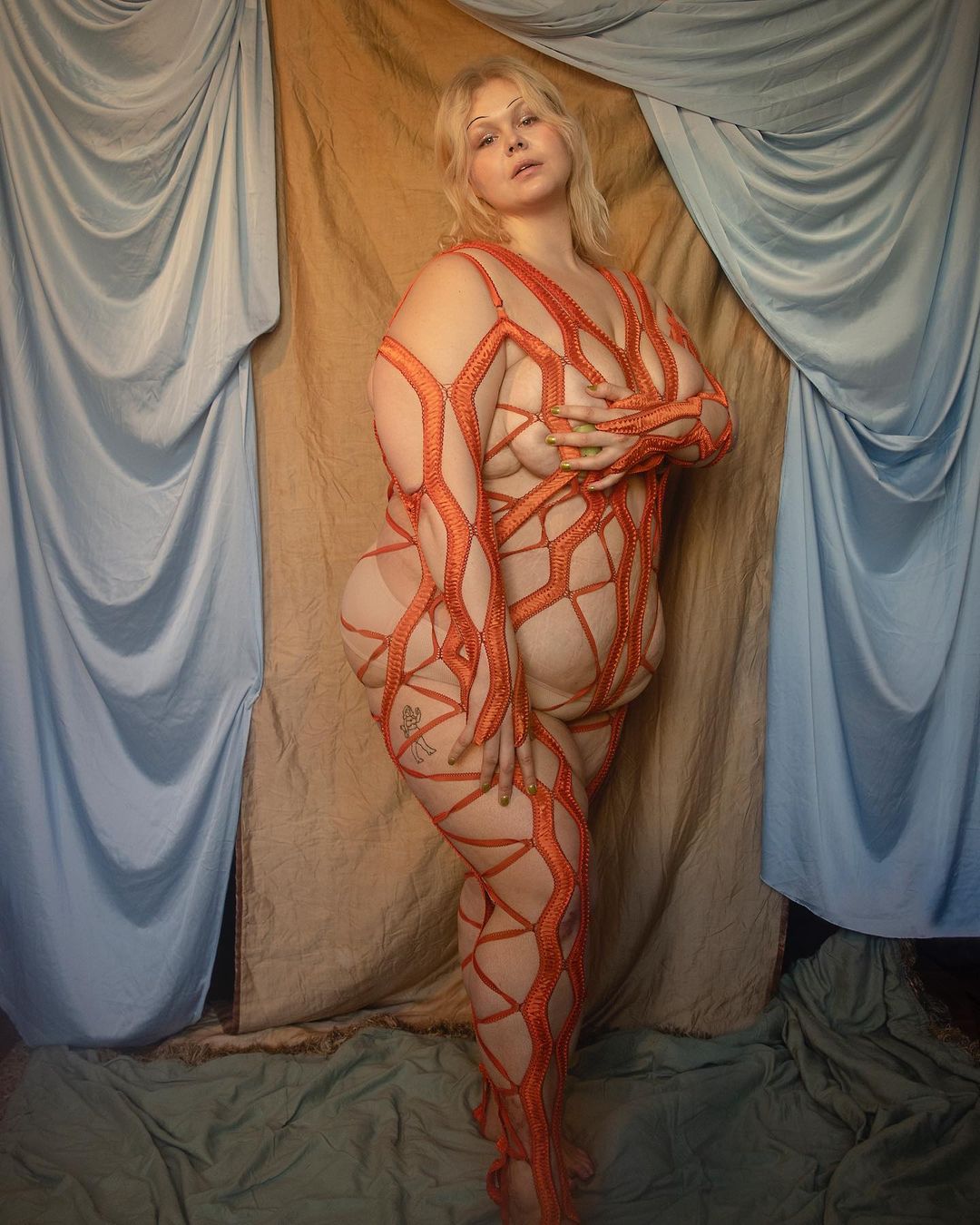 Adult woman poses in body stockings in front of draped fabric backdrop with soft lighting, creating a fine art portrait.