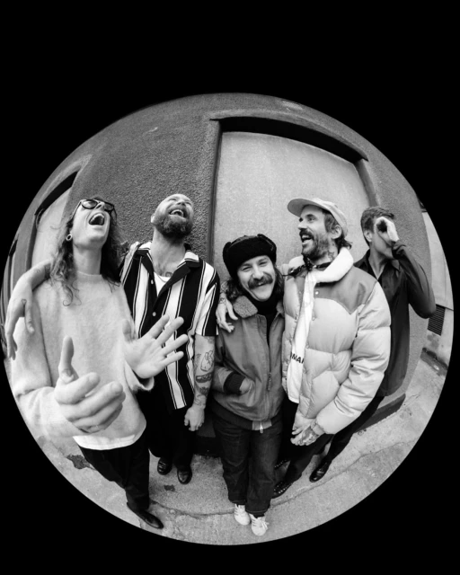 Black and white portrait of a band outdoors, featuring five members in varied expressions, fisheye lens, and urban background.