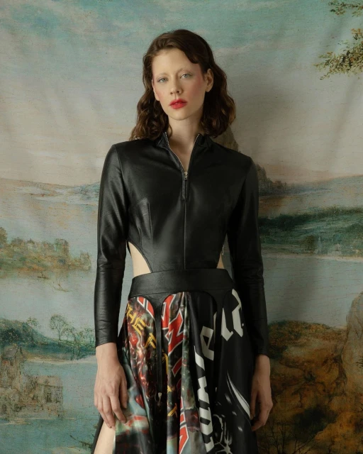 A woman with red lipstick poses in a black top and printed skirt against a painterly backdrop in a fashion studio portrait.