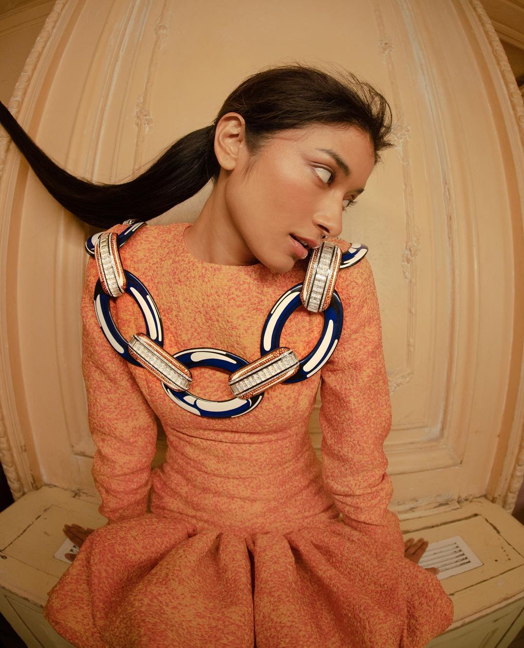 Stylish woman in orange dress with bold chain necklace poses in an indoor setting with neutral backdrop. Fashion editorial portrait.