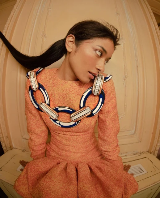 Stylish woman in orange dress with bold chain necklace poses in an indoor setting with neutral backdrop. Fashion editorial portrait.