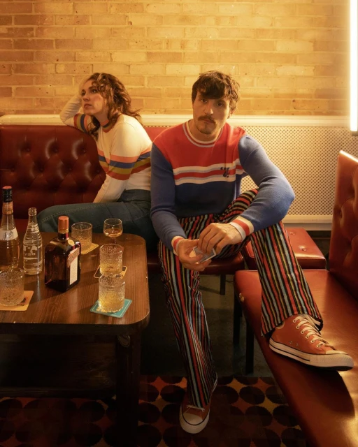 Two people sitting in a bar. A man wearing striped pants and a colorful sweater and a woman wearing a striped sweater.