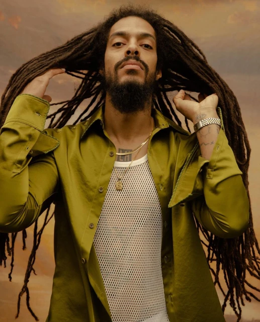 Portrait of a man with long dreadlocks, wearing a light-green collared shirt and white fishnet undershirt, against a warm-toned background.