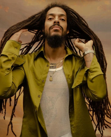 Portrait of a man with long dreadlocks, wearing a light-green collared shirt and white fishnet undershirt, against a warm-toned background.