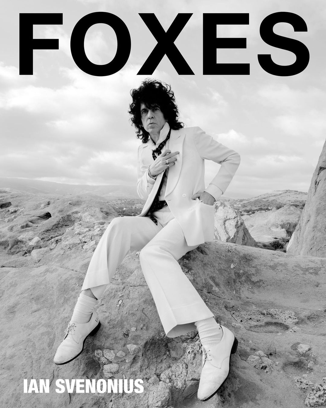 Monochrome portrait of Ian Svenonius in a white suit, seated outdoors against a rocky, desert-like backdrop, with the text 'FOXES' above.