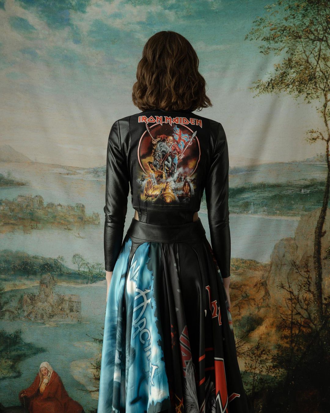 Woman seen from behind wearing an Iron Maiden printed leather jacket and graphic long skirt against a landscape tapestry backdrop.