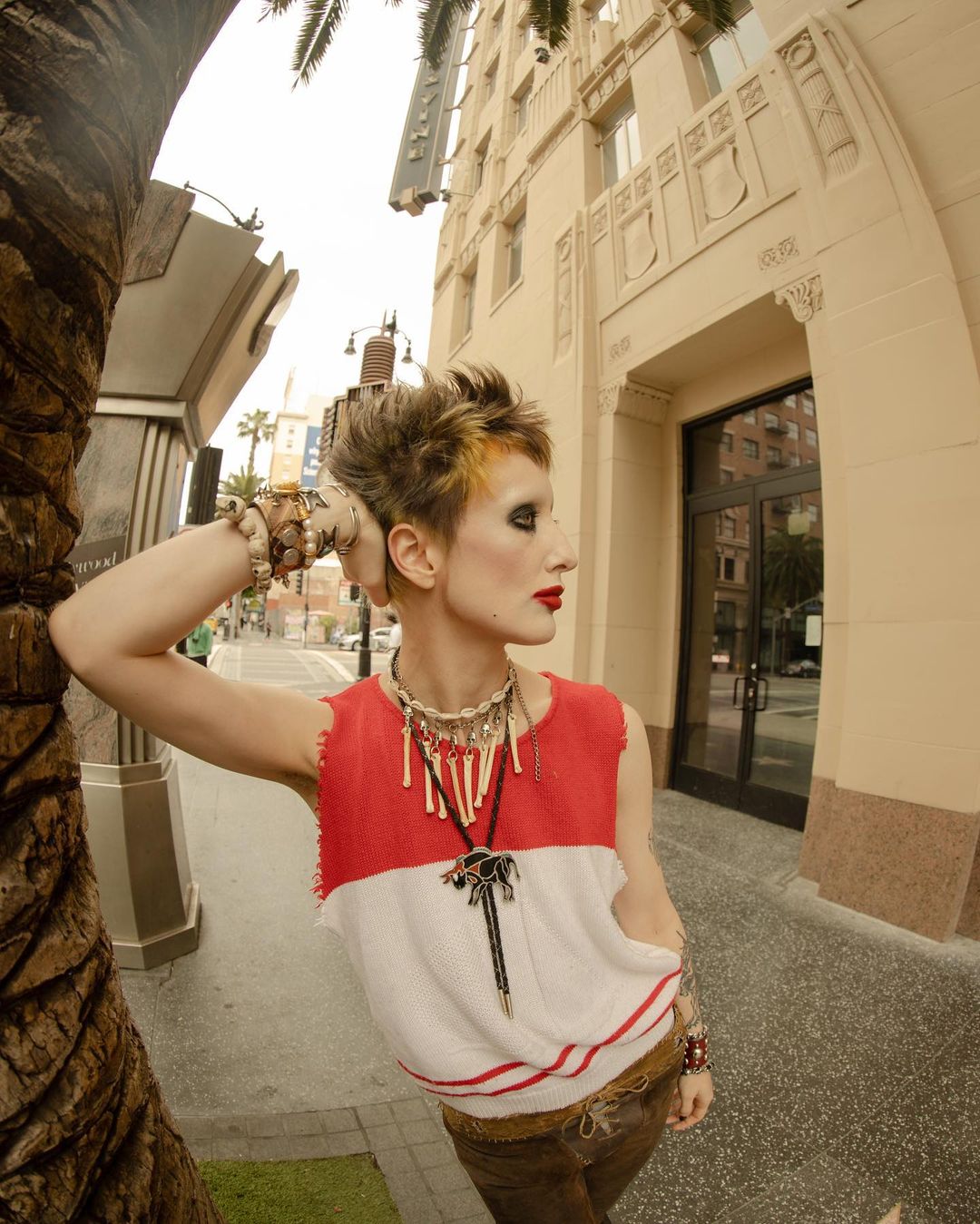 Wide-angle shot of an adult female in a red and white top posing outdoors next to a palm tree and building with architectural details.