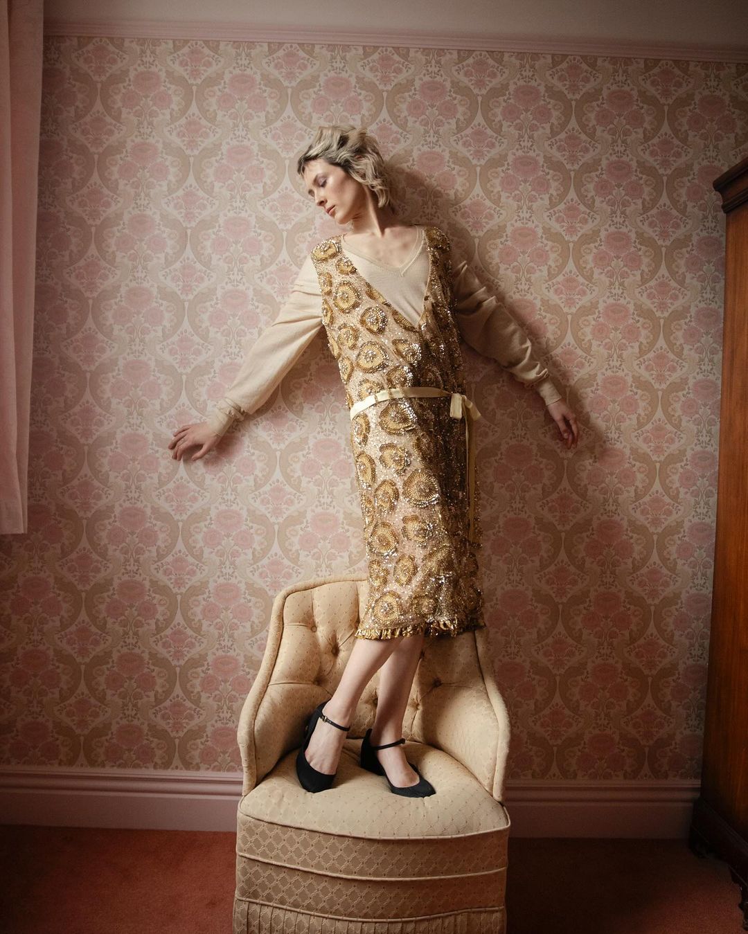 Adult female in gold flapper dress posing on chair in front of wallpaper in an indoor setting, looking down in a three-quarter shot.