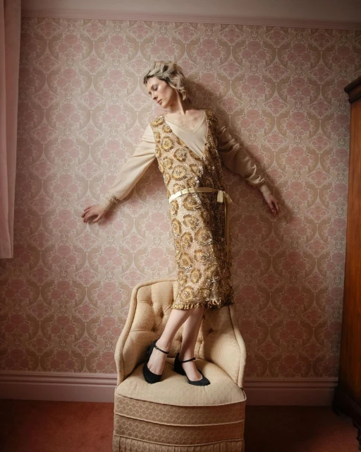 Adult female in gold flapper dress posing on chair in front of wallpaper in an indoor setting, looking down in a three-quarter shot.
