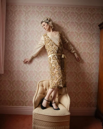 Adult female in gold flapper dress posing on chair in front of wallpaper in an indoor setting, looking down in a three-quarter shot.