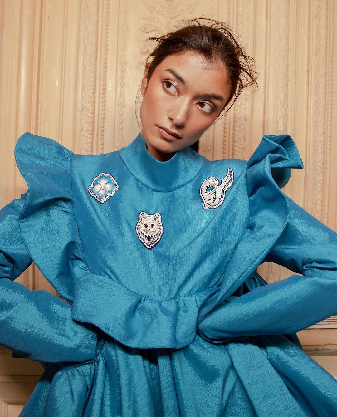 Close-up of woman in a blue ruffled dress with decorative patches. She looks away from the camera with a slight smile in a bright, indoor setting.
