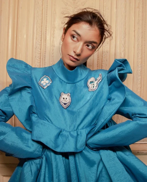 Close-up of woman in a blue ruffled dress with decorative patches. She looks away from the camera with a slight smile in a bright, indoor setting.