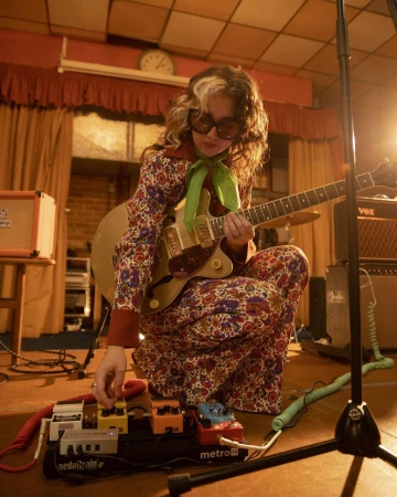 Woman plays electric guitar on stage in a floral pantsuit with vintage amps and pedal board equipment under warm performance lighting.