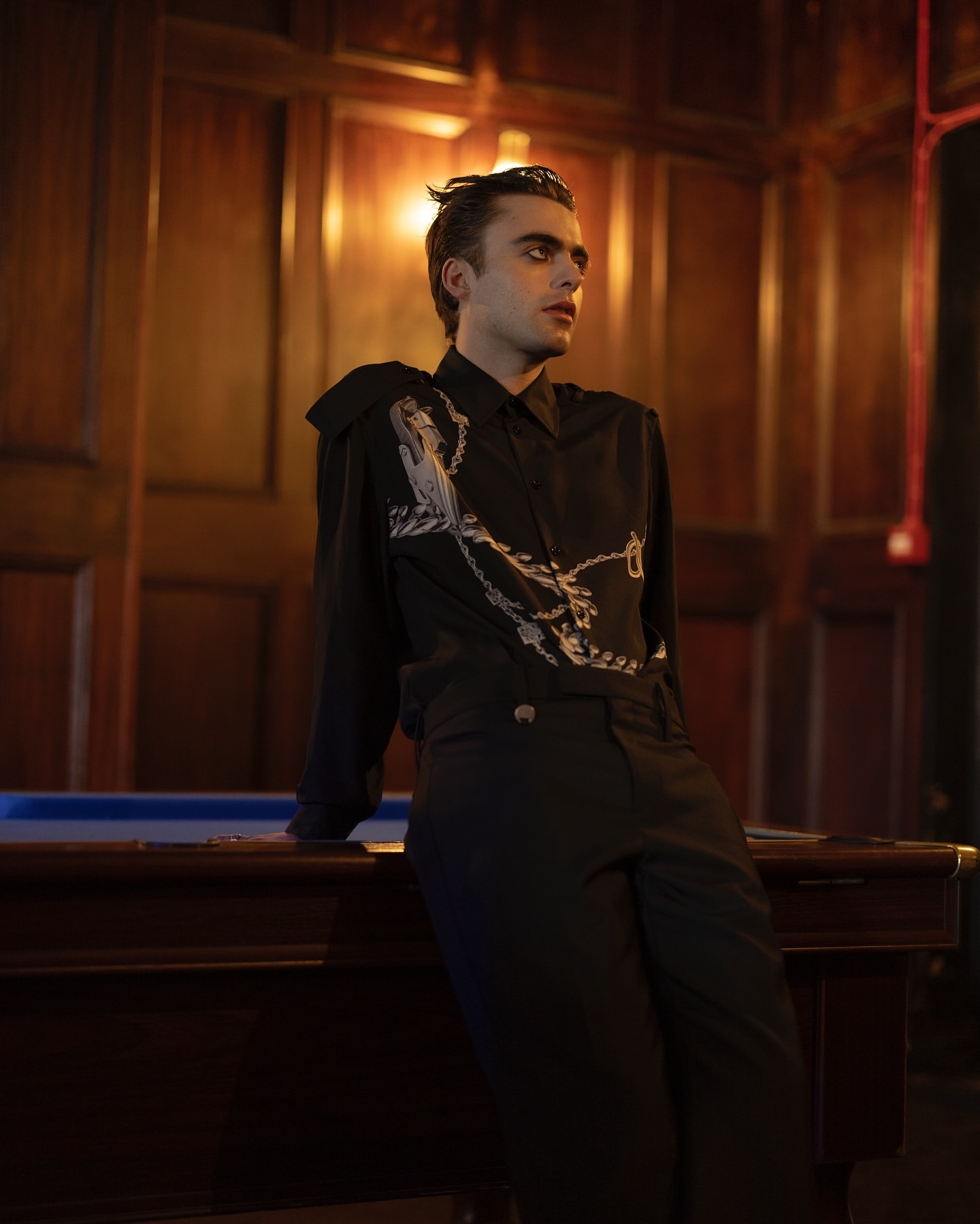 Stylish young man with dark hair and dramatic eye makeup posing near a pool table indoors with dark wood paneling.