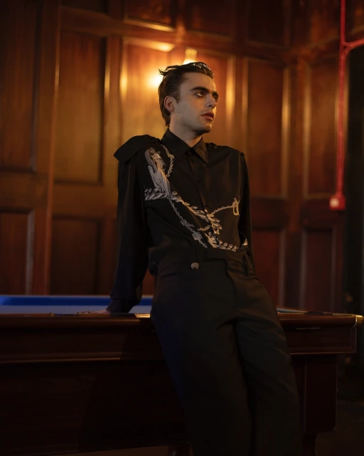 Stylish young man with dark hair and dramatic eye makeup posing near a pool table indoors with dark wood paneling.