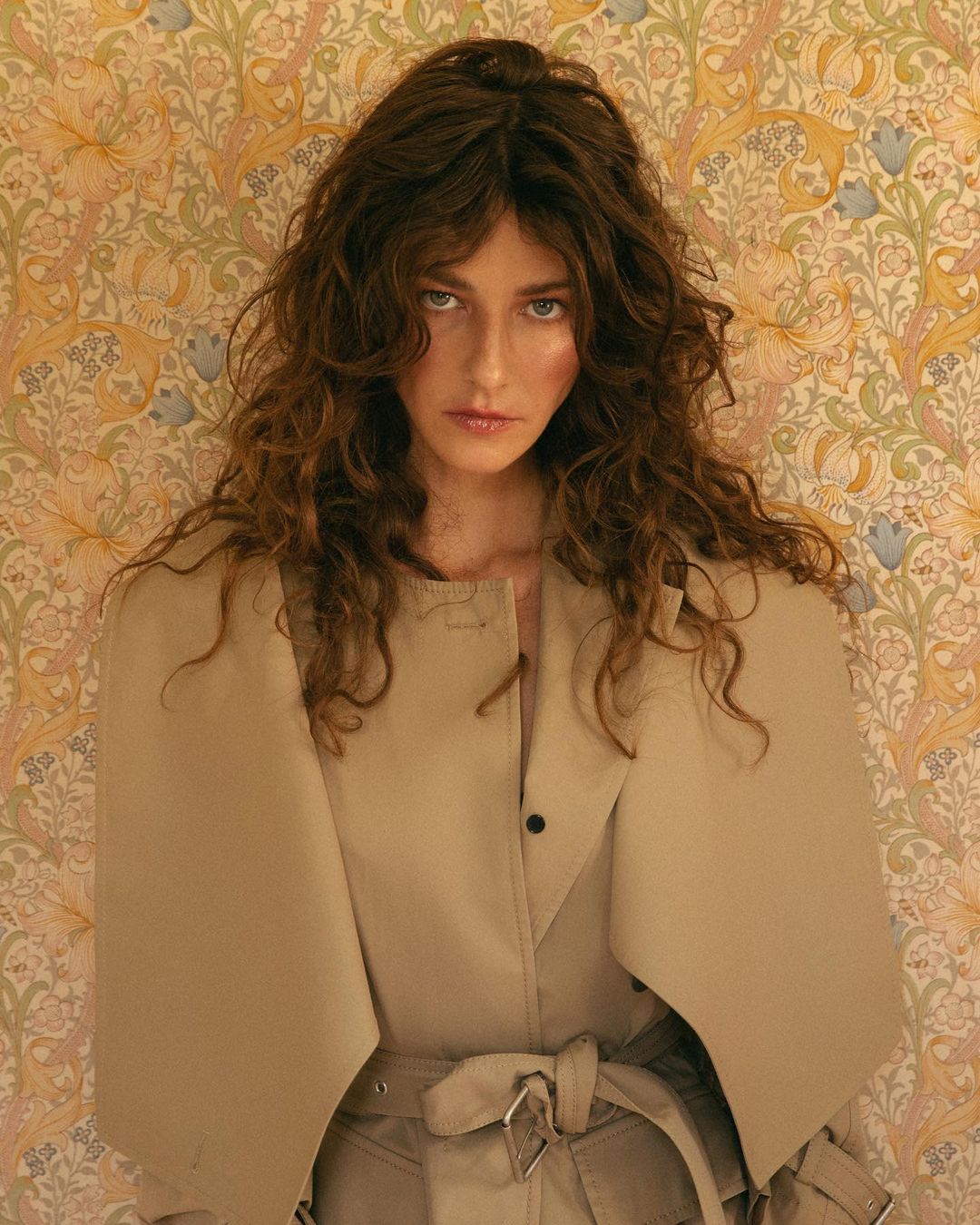 Indoor portrait of an adult woman with curly brown hair wearing a tan trench coat, posing in front of a vintage floral wallpaper.